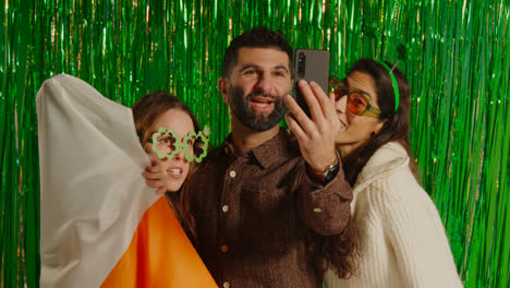 Studio-Shot-Of-Friends-Dressing-Up-With-Irish-Novelties-And-Props-Posing-For-Selfie-Celebrating-St-Patrick's-Day-Against-Green-Tinsel-Background-3